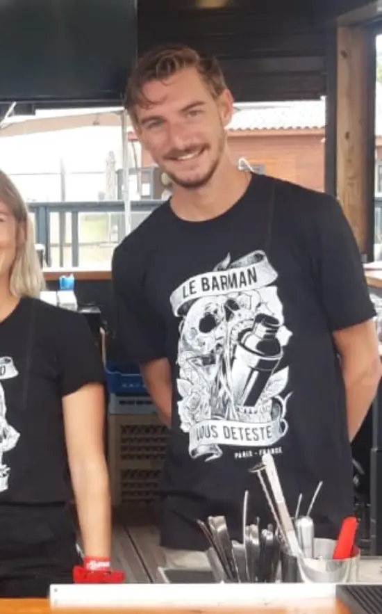 une photo d'un homme dérière un bar, avec un teeshirt noir où il est écrit :"Le barman vous déteste"